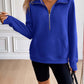 Woman wearing Ivy Lane Half Zip Raglan Sleeve Sweatshirt in blue with front pocket, standing in a stylish room with wooden floor and white wall paneling.