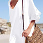 Woman wearing a white ruffled v-neck cover up at the beach, stylishly holding a woven bag.