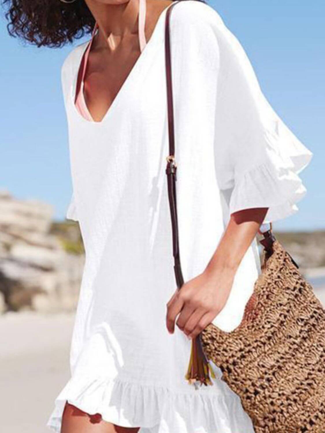 Woman wearing a white ruffled v-neck cover up at the beach, stylishly holding a woven bag.