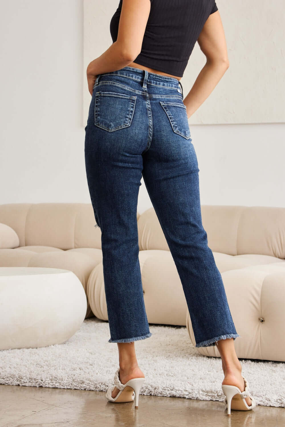 Woman wearing Crop Dylan Full Size Tummy Control Distressed High Waist Raw Hem Jeans from RFM Jeans, showcasing back view.