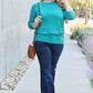 Woman wearing a teal round neck batwing sleeve top with jeans, carrying a brown shoulder bag, and smiling while adjusting sunglasses.