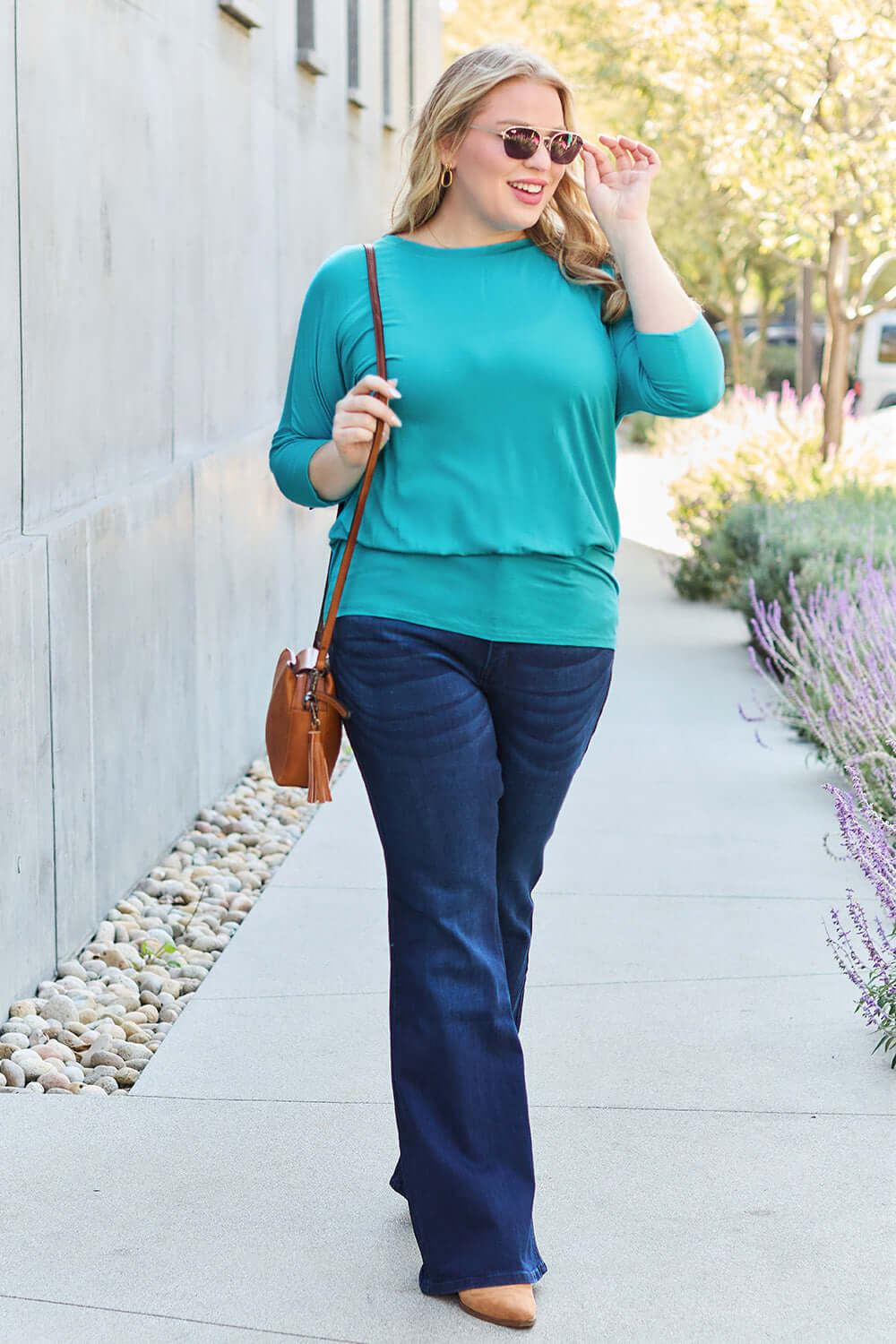 Woman wearing a teal round neck batwing sleeve top with jeans, carrying a brown shoulder bag, and smiling while adjusting sunglasses.