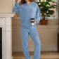 Woman in light blue Bella Road hoodie and pants set holding coffee, standing by a fireplace. Cozy and stylish two-piece outfit.