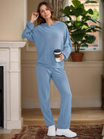 Woman in light blue Bella Road hoodie and pants set holding coffee, standing by a fireplace. Cozy and stylish two-piece outfit.