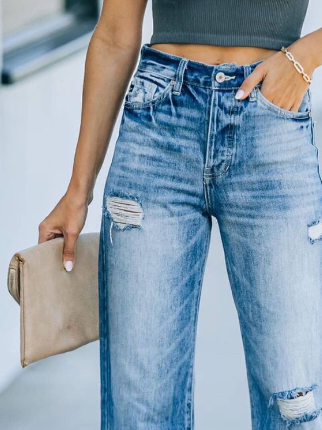 Woman wearing distressed straight leg jeans with hand in pocket, holding a beige clutch bag.