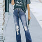 Woman wearing distressed bootcut jeans with pockets, graphic tee, and sandals, standing on street with pavement background.