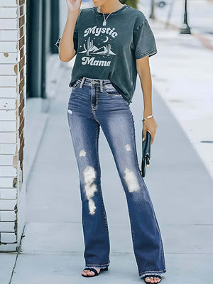 Woman wearing distressed bootcut jeans with pockets, graphic tee, and sandals, standing on street with pavement background.