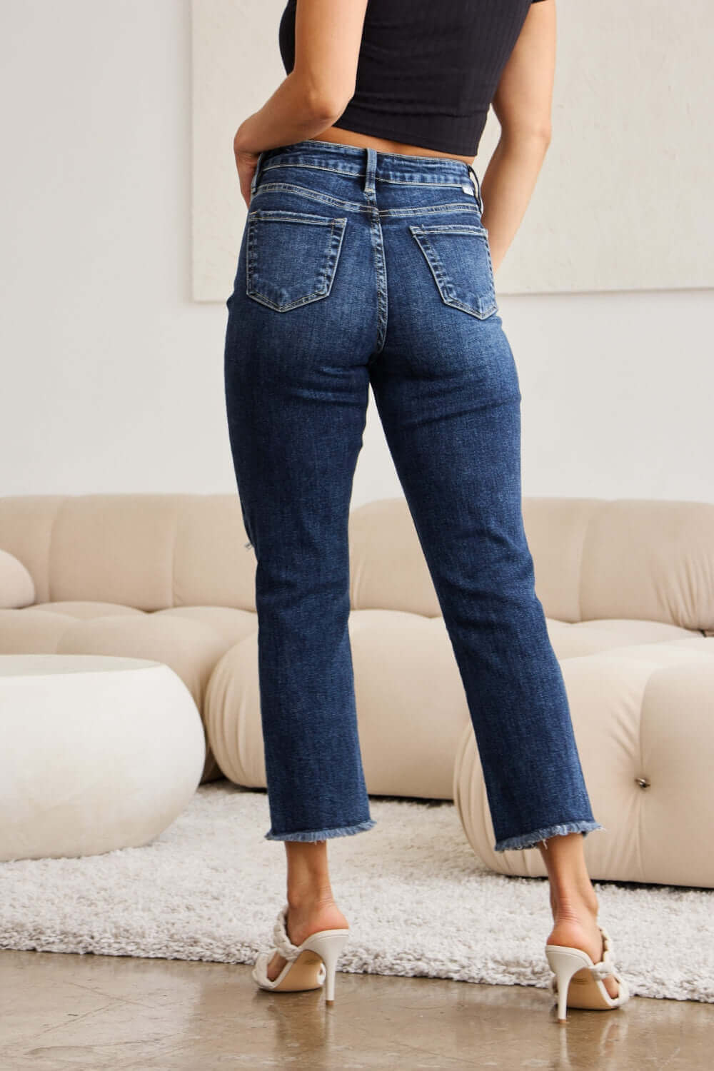 Woman wearing RFM Jeans Crop Dylan Full Size Tummy Control Distressed High Waist Raw Hem Jeans standing in a modern living room.