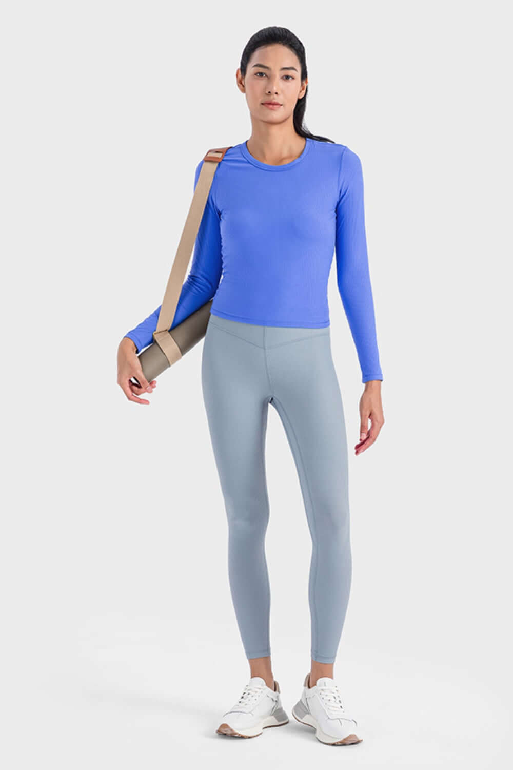 Woman in blue long sleeve sports top and gray leggings, ready for a workout with a yoga mat bag.