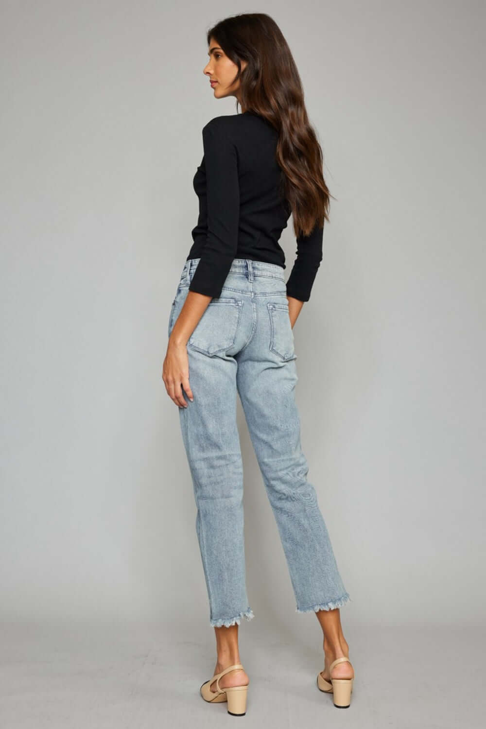 Woman wearing high waist button fly raw hem cropped straight jeans with black top and beige sandals, showcasing the back view.