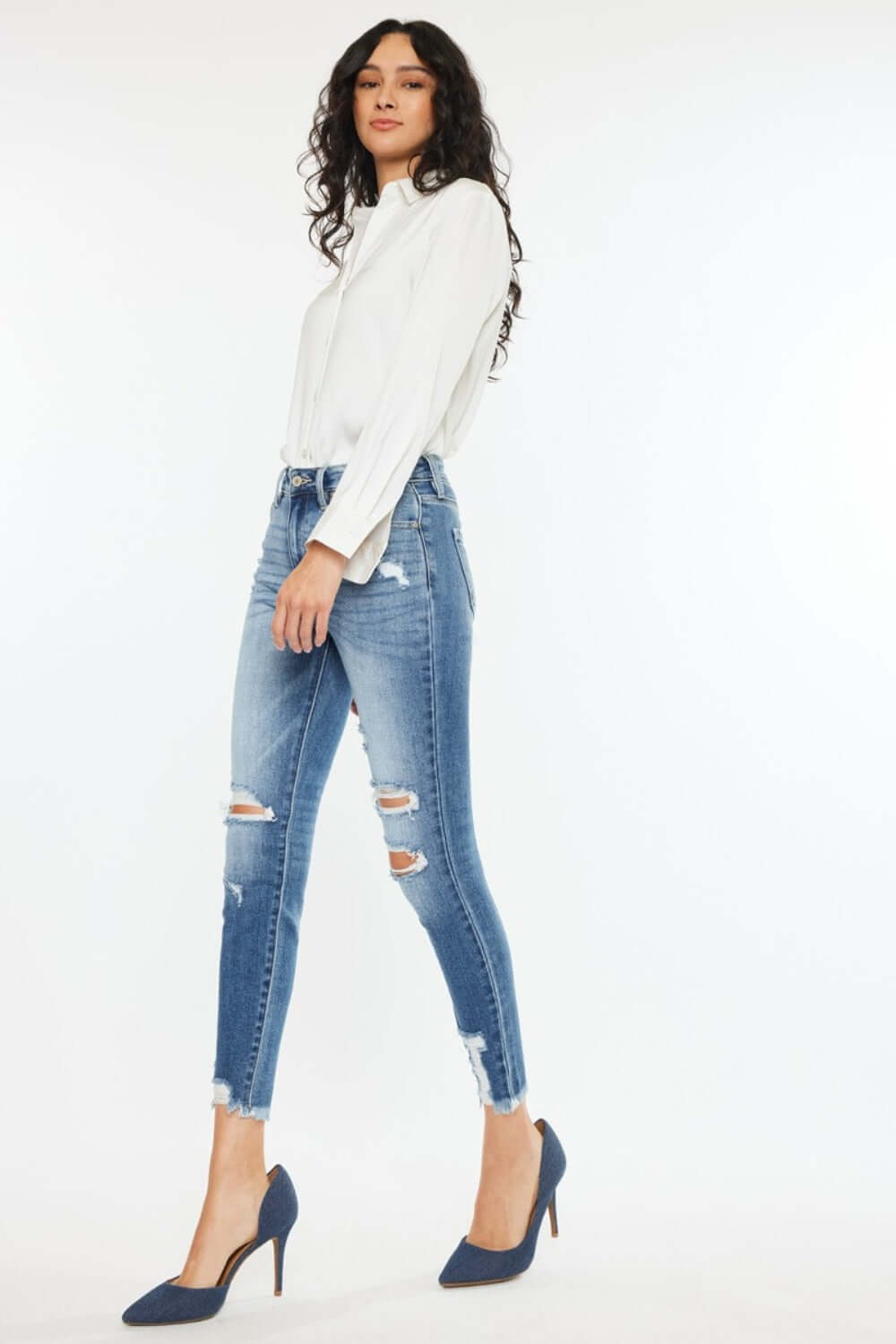 Woman wearing high rise frayed ankle skinny jeans in medium wash with distressed details and fray hem, paired with white blouse and heels