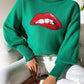 Woman wearing Bella Road Lip Turtleneck Long Sleeve Sweater in green, featuring bold red lips design, sitting cross-legged in white pants.