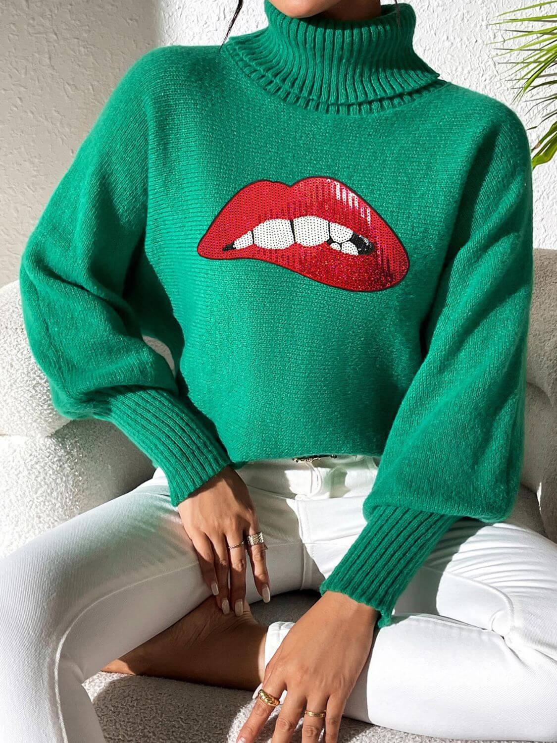 Woman wearing Bella Road Lip Turtleneck Long Sleeve Sweater in green, featuring bold red lips design, sitting cross-legged in white pants.