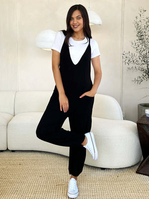 Woman wearing a black plunge sleeveless jumpsuit with pockets, white shirt, and white sneakers, posing confidently in a stylish room.