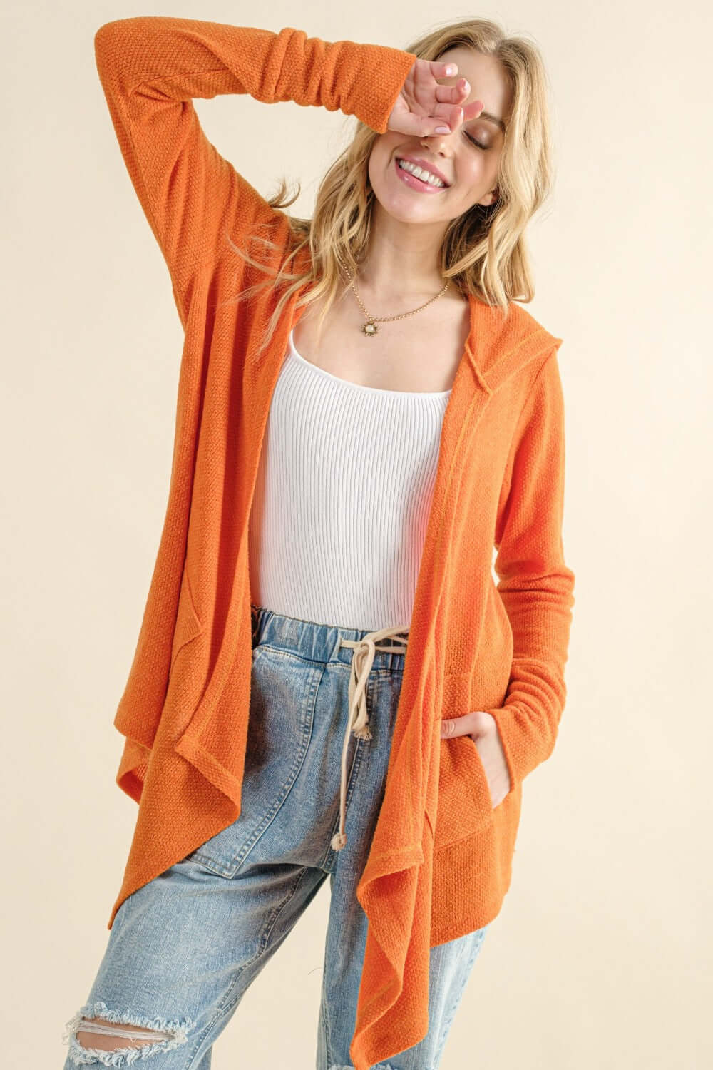 Woman wearing orange thermal hooded open front cardigan with pockets, styled with a white tank top and ripped jeans, looking cozy and fashionable