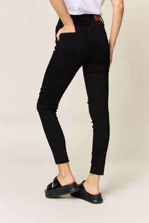 Rear view of woman wearing distressed tummy control high waist skinny jeans by Judy Blue Jeans in black, featuring trendy raw hems.