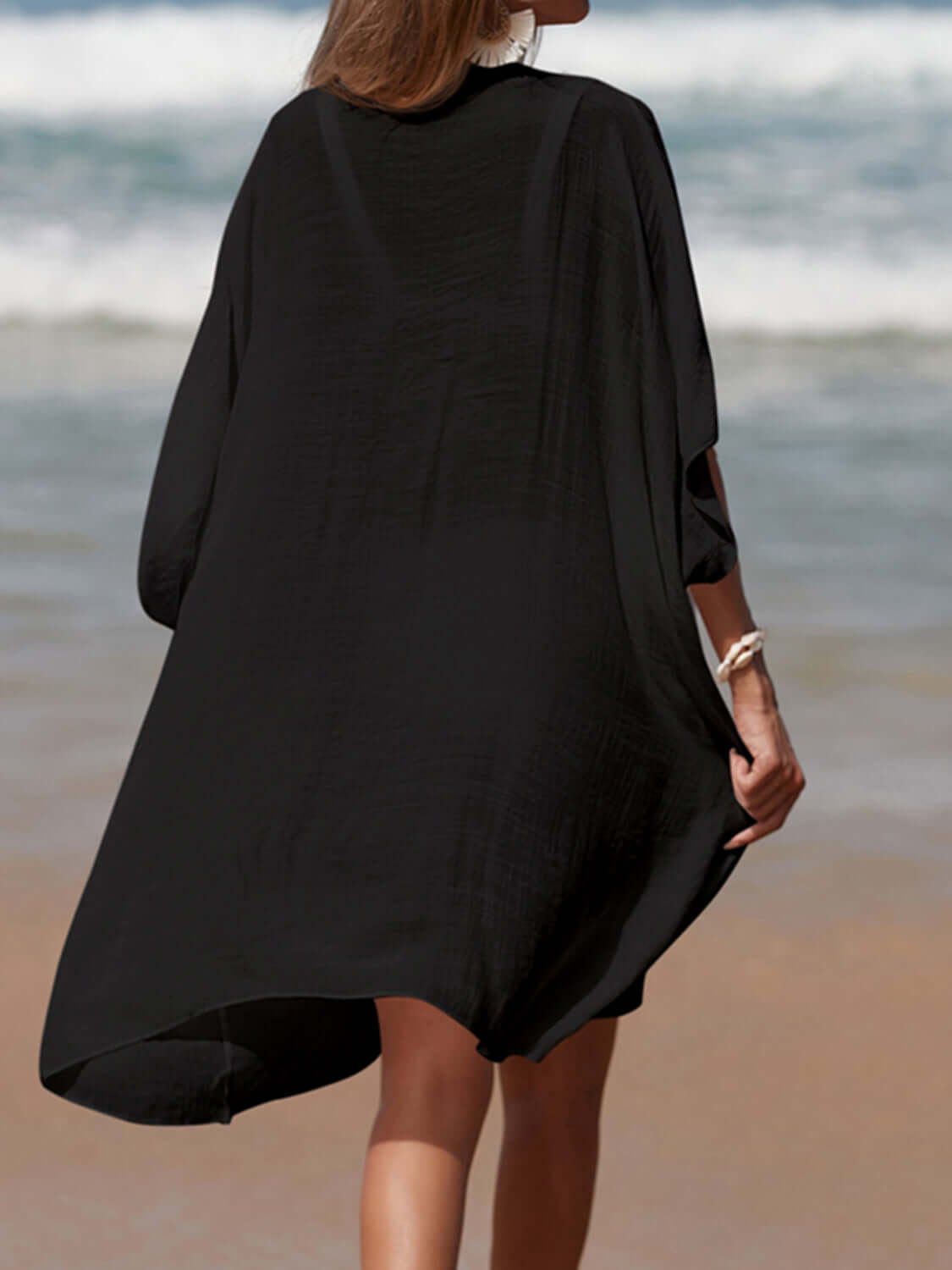 Woman in black cutout V-neck three-quarter sleeve cover up walking on beach. Openwork sheer fabric, no stretch. Cotton-polyester blend.