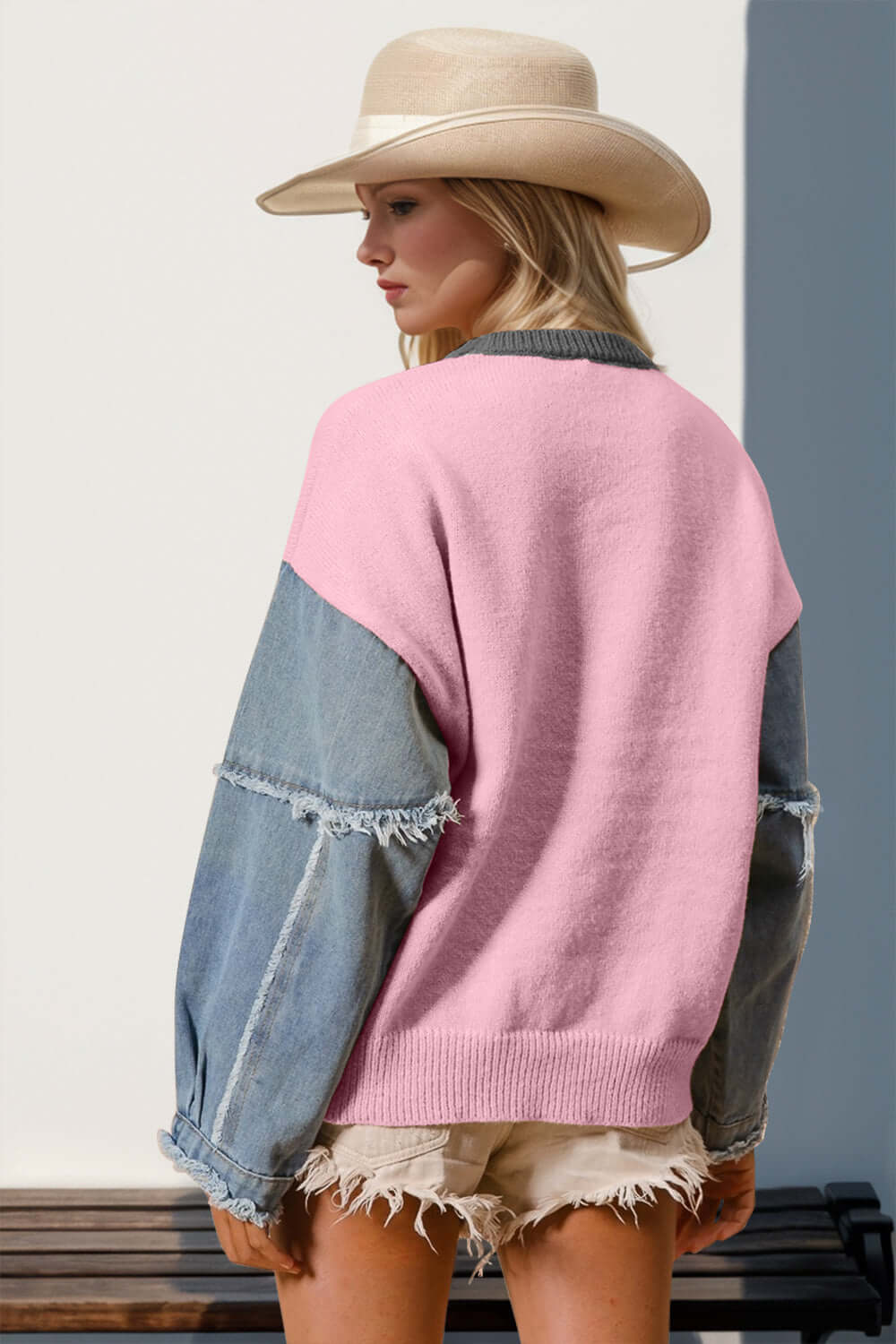 Woman in pink sweater with raw edge denim sleeves, wearing a hat and shorts, stands stylishly outdoors.