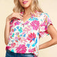 Woman wearing notch neck sequin floral blouse with vibrant pink and blue flower patterns, paired with casual blue jeans.