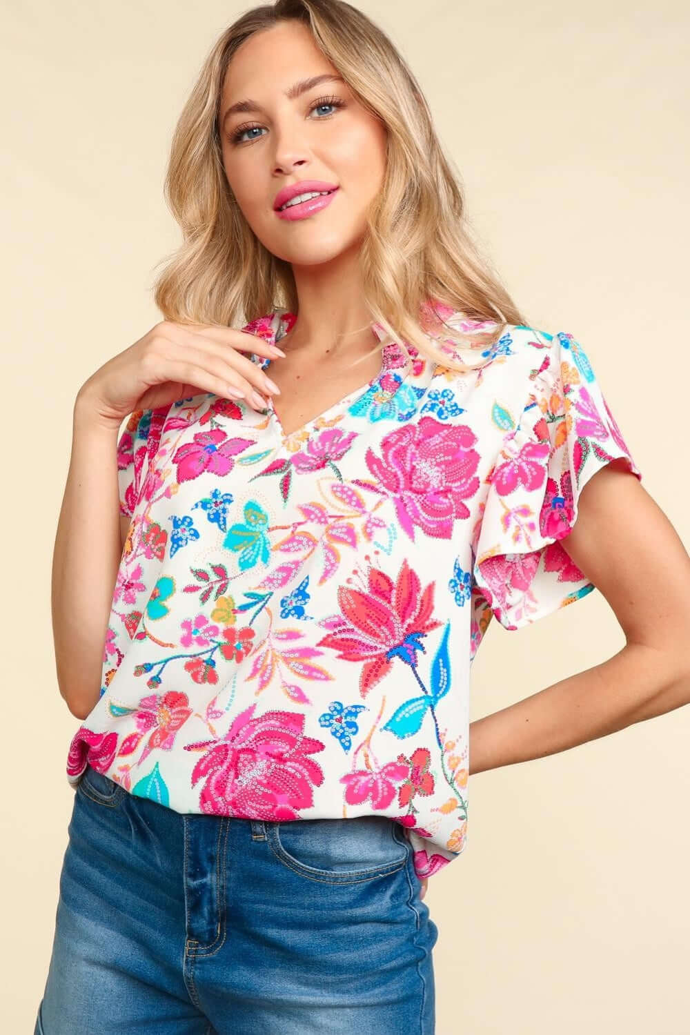 Woman wearing notch neck sequin floral blouse with vibrant pink and blue flower patterns, paired with casual blue jeans.