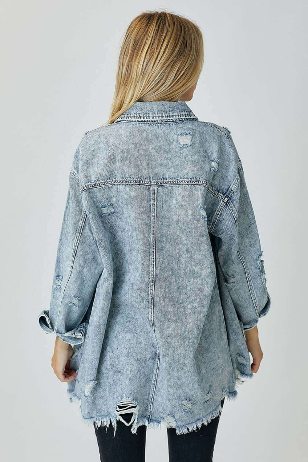 Back view of a distressed raw hem denim shirt by Risen Jeans, featuring frayed edges and a relaxed fit for a casual, rugged look.