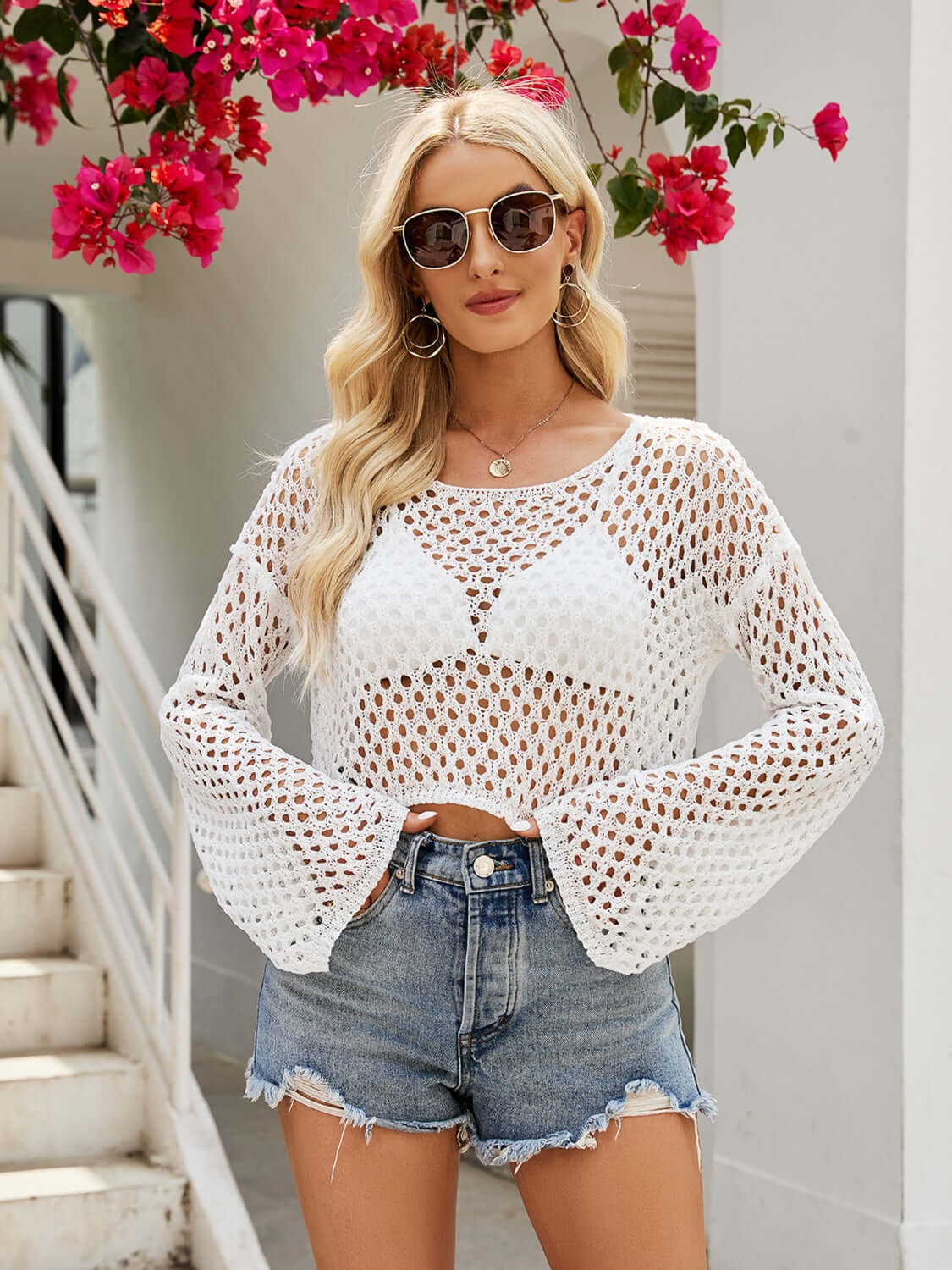 Woman wearing openwork round neck long sleeve cover-up with denim shorts and sunglasses, standing near stairs with pink flowers in the background