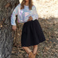 Woman wearing J.NNA sequin long sleeve crop t-shirt, black skirt, and cowboy boots, leaning against a tree outdoors.