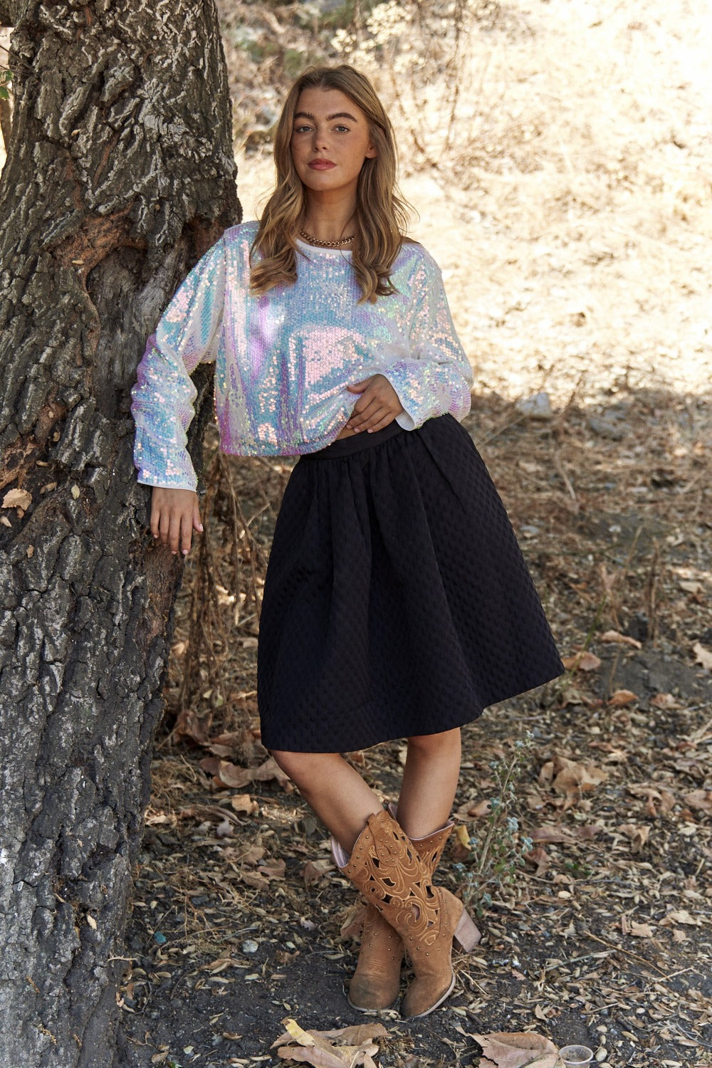 Woman wearing J.NNA sequin long sleeve crop t-shirt, black skirt, and cowboy boots, leaning against a tree outdoors.