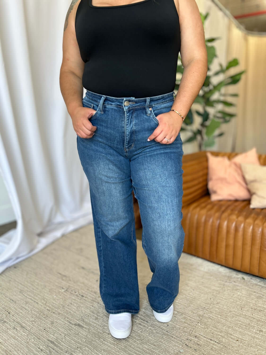 Woman wearing RFM full-size high-rise tummy control wide leg jeans with a black top and white sneakers in a modern living room.