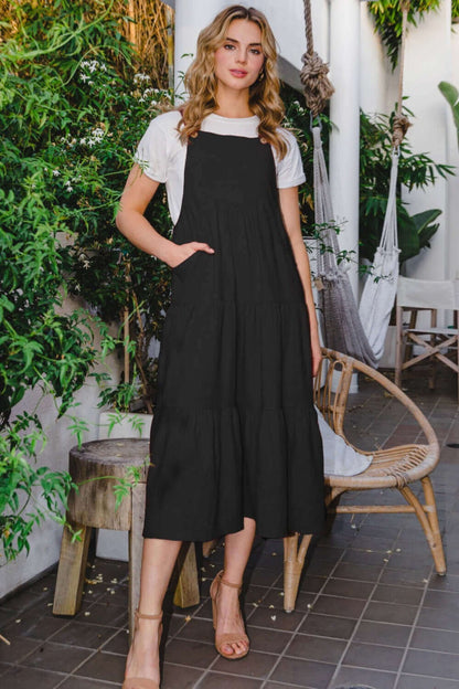 Woman wearing Sleeveless Tiered Midi Dress in black, posing outdoors amidst lush greenery and patio furniture.