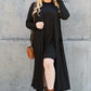 Woman in open front long sleeve cover up, black dress, and brown boots standing against a concrete wall