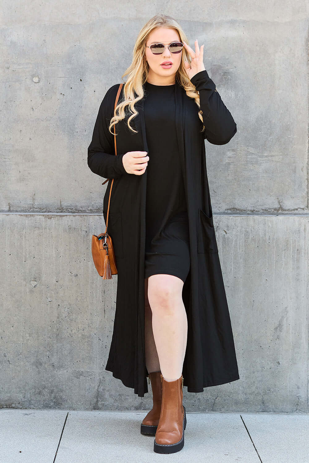 Woman in open front long sleeve cover up, black dress, and brown boots standing against a concrete wall