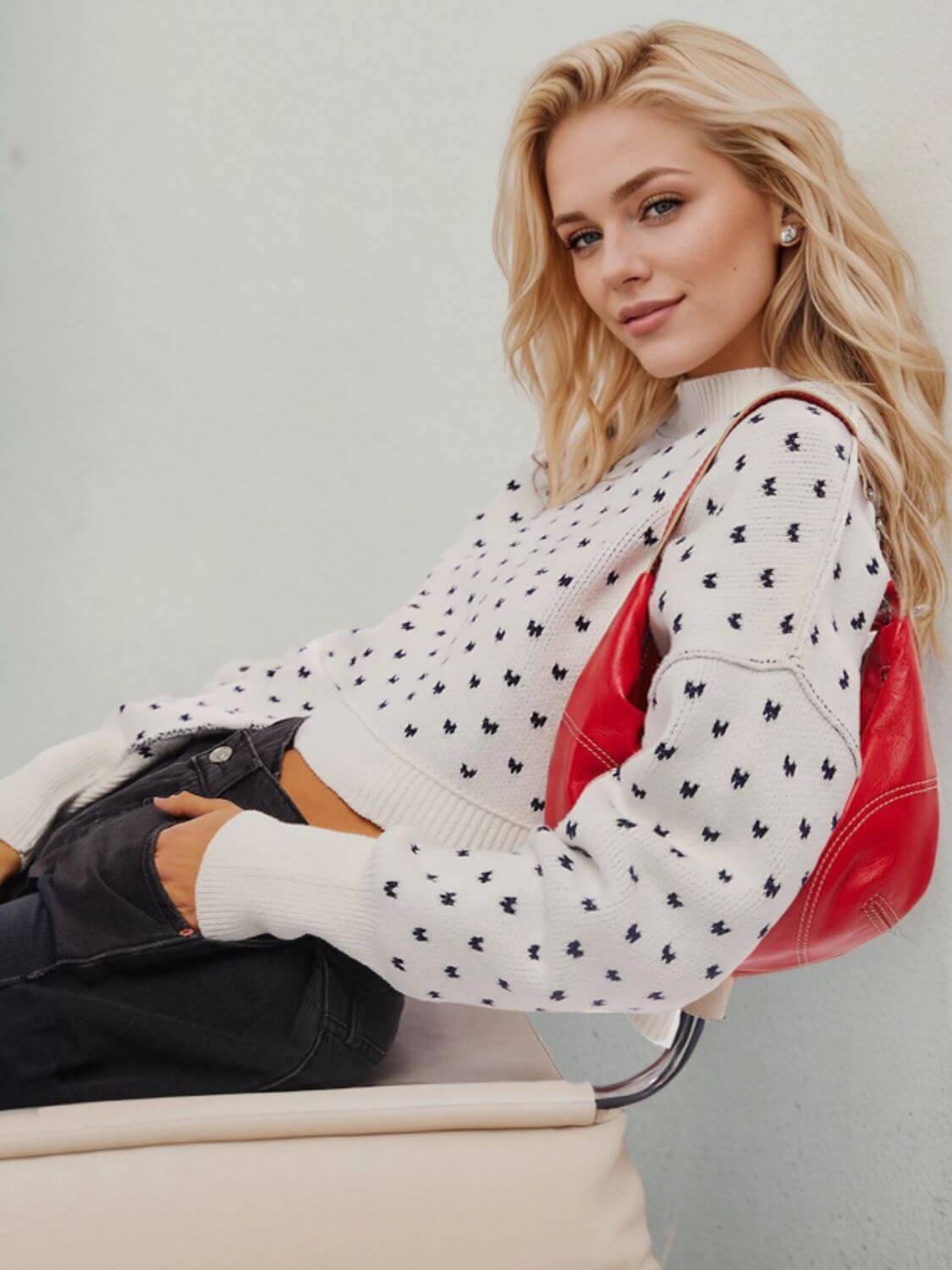 Stylish woman wearing a cozy white sweater with black patterns and holding a red bag, seated comfortably.