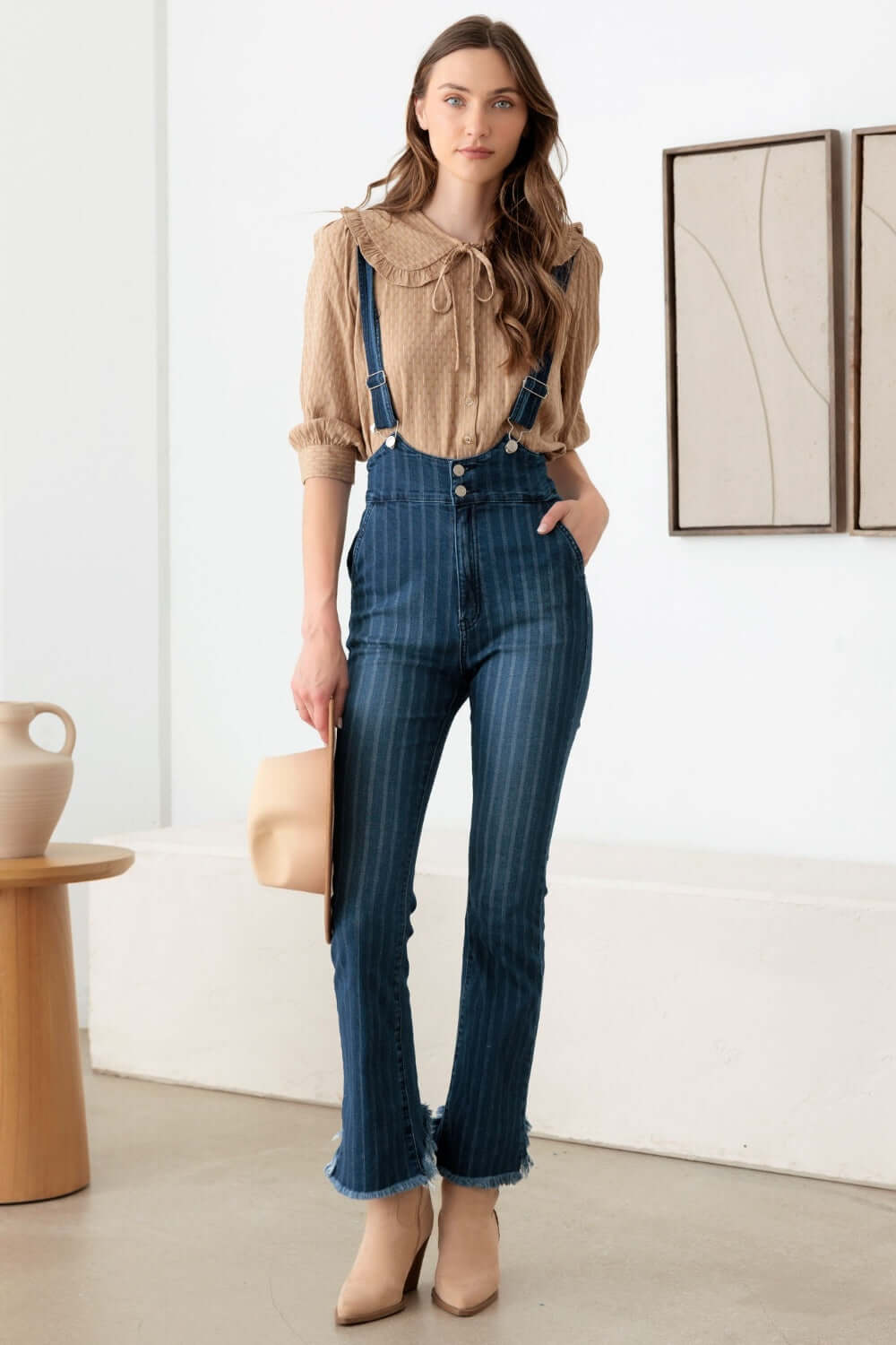 Woman wearing striped stretched suspender denim overalls with high waist, front pockets, front zip, and destroyed hem for a retro-inspired look