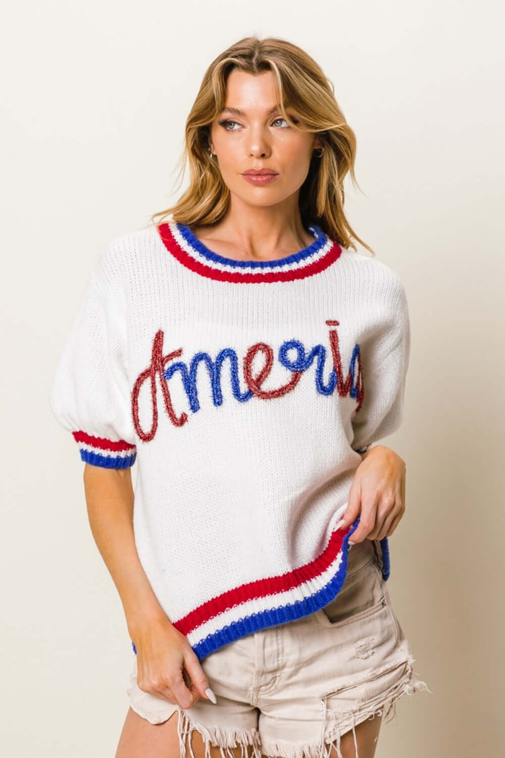 Woman wearing a metallic America letter short sleeve sweater with patriotic red, white, and blue detailing, paired with beige shorts.