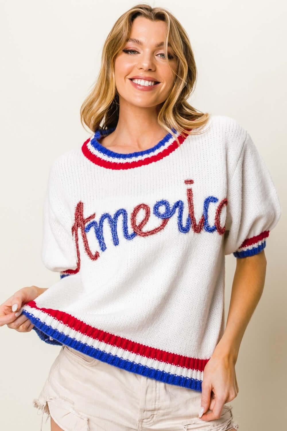 Woman wearing a metallic America letter short sleeve sweater with red, white, and blue detailing. Trendy and patriotic fashion.