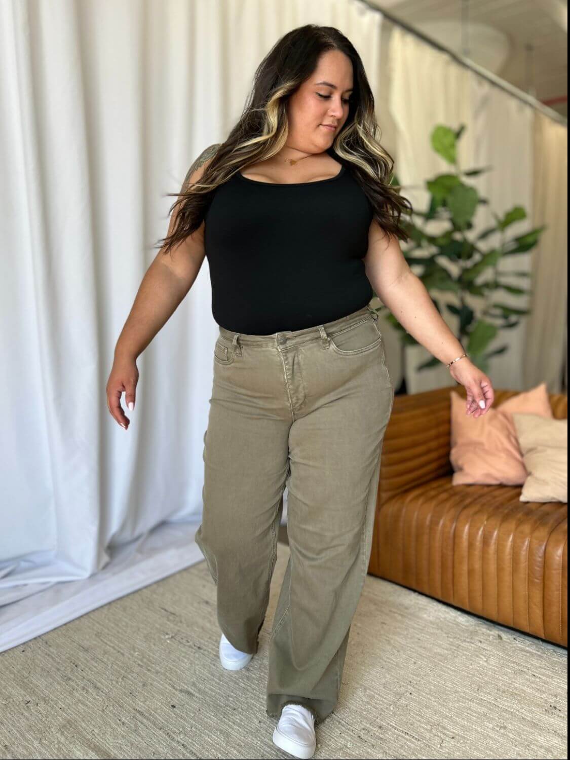 Woman wearing high-rise wide-leg jeans and a black top stands confidently in a modern, cozy room with white curtains and a brown sofa.