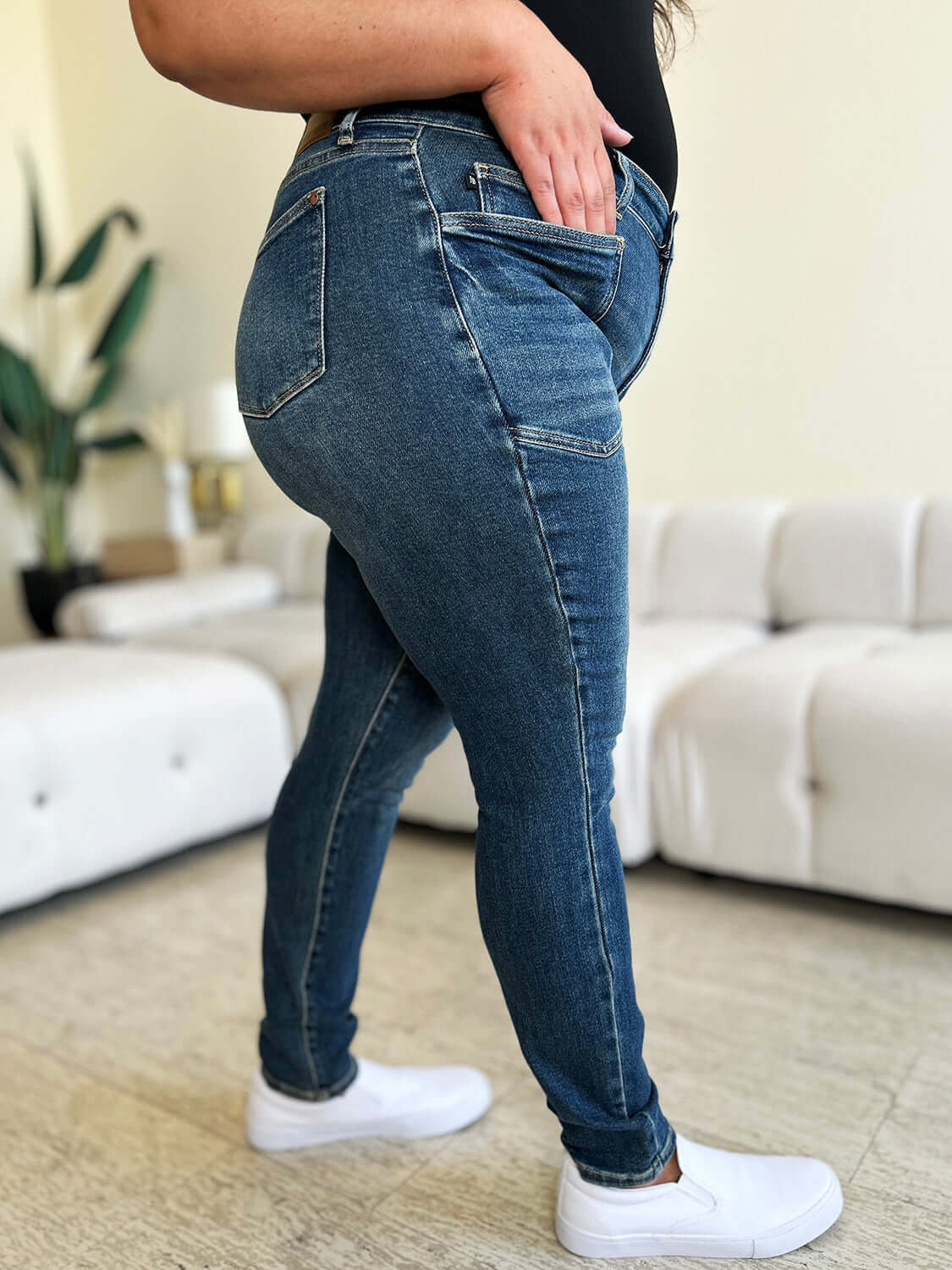 Woman wearing Judy Blue High Waist Skinny Jeans in living room with white sneakers and black top.