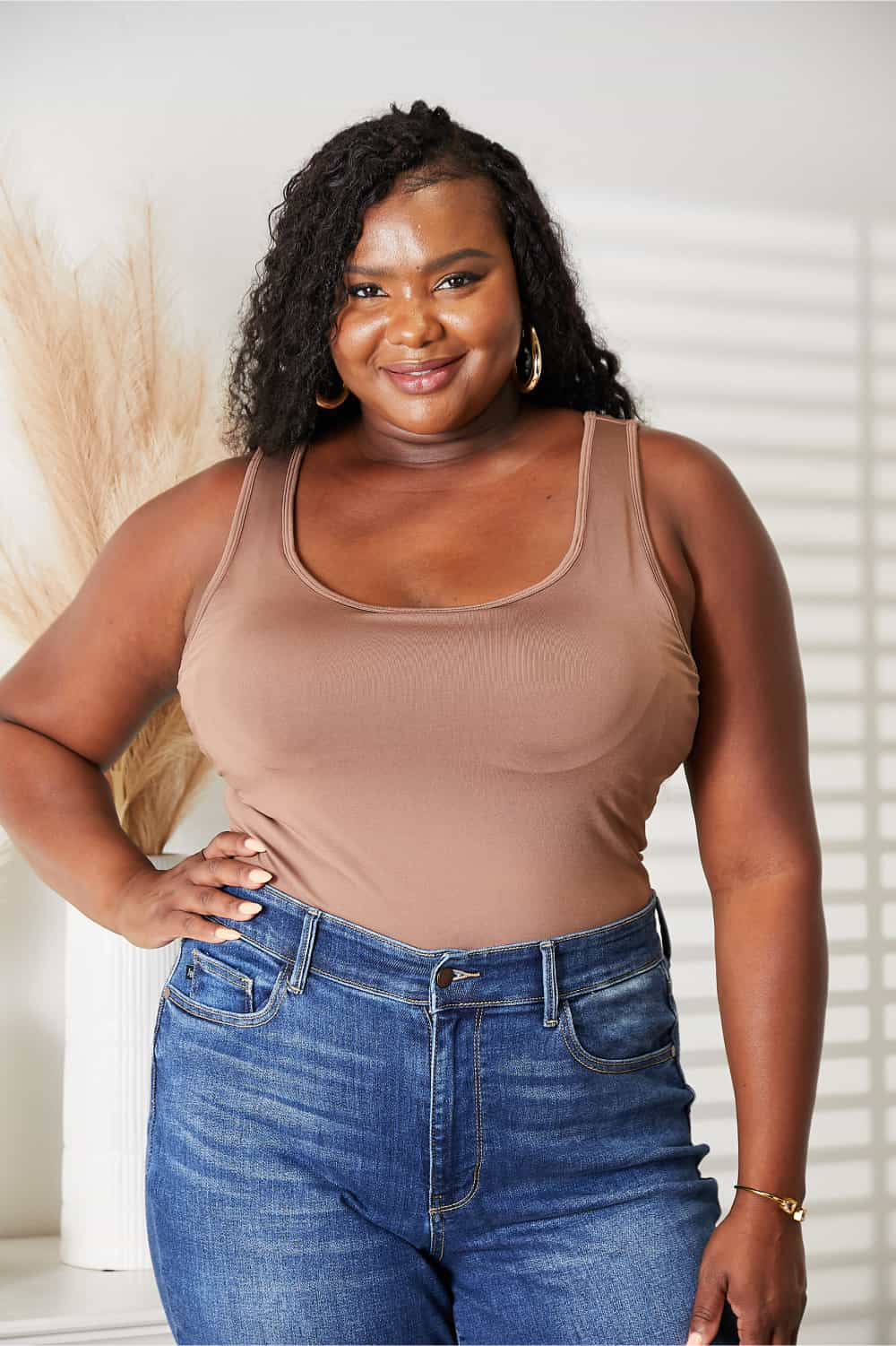 Woman wearing everyday full-size basic tank bodysuit paired with blue jeans, with a backdrop of neutral decor.