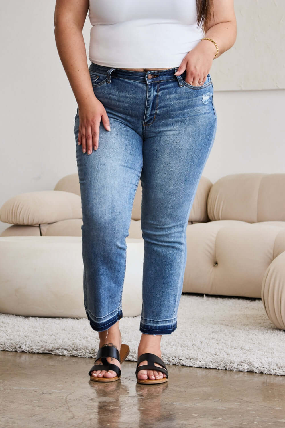 Woman wearing Judy Blue release hem cropped bootcut jeans with sandals in living room.