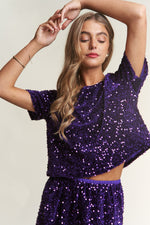 Woman wearing a J.NNA sequin short sleeve crop T-shirt in purple, highlighting its dazzling sparkle and stylish cropped design.