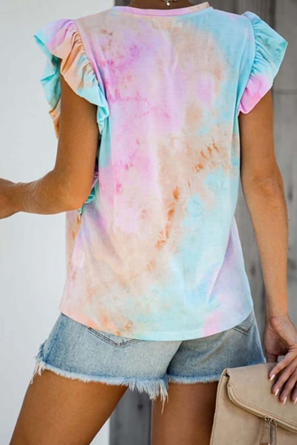 Woman wearing Bella Road Ruffled Printed T-Shirt in pastel tie-dye, round neck, cap sleeves, with denim shorts and a beige bag.