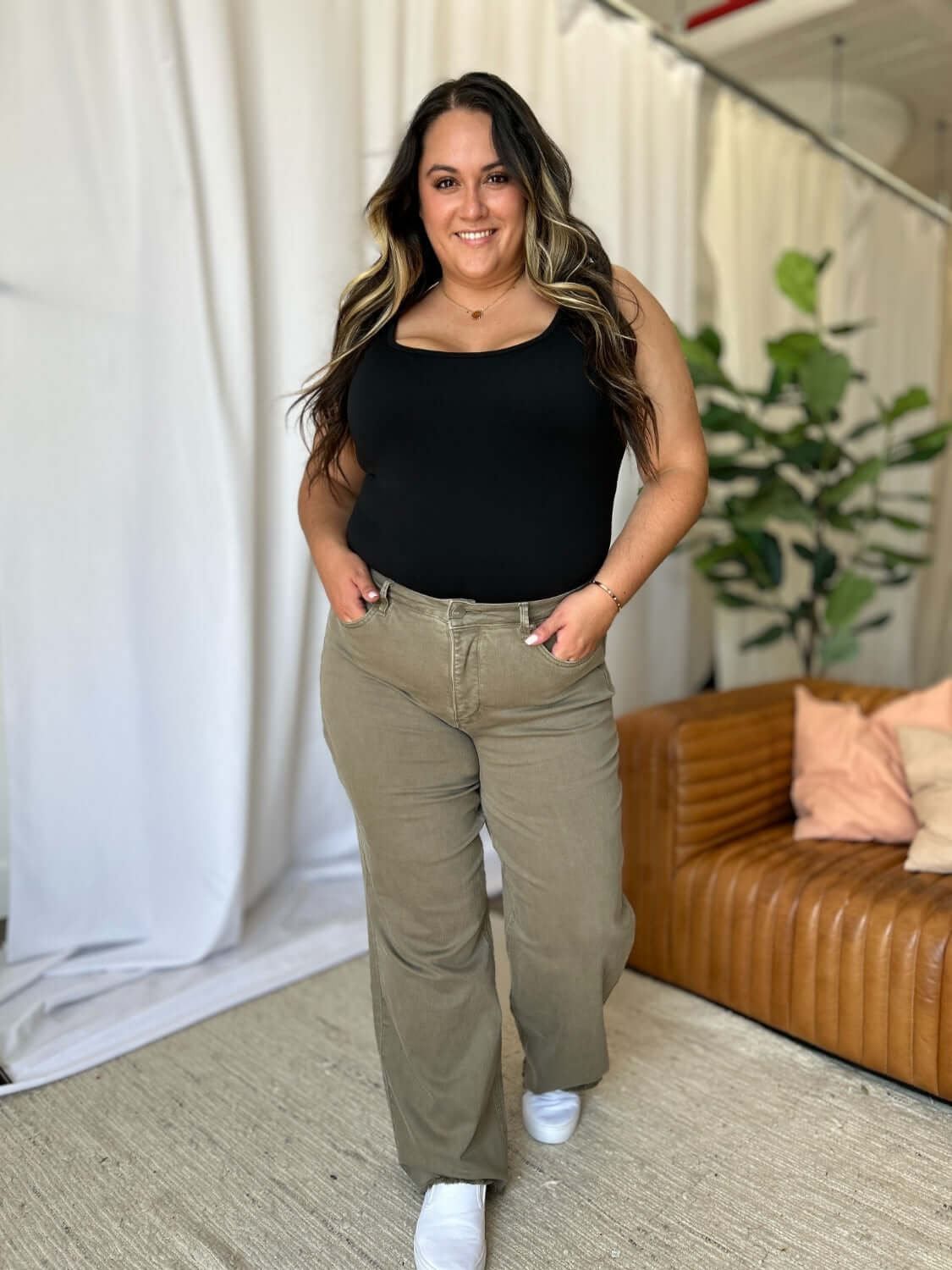 Woman wearing RFM Full Size High Rise Garment Dye Wide Leg Jeans with a black top, standing in a stylish living room with plants and a brown sofa