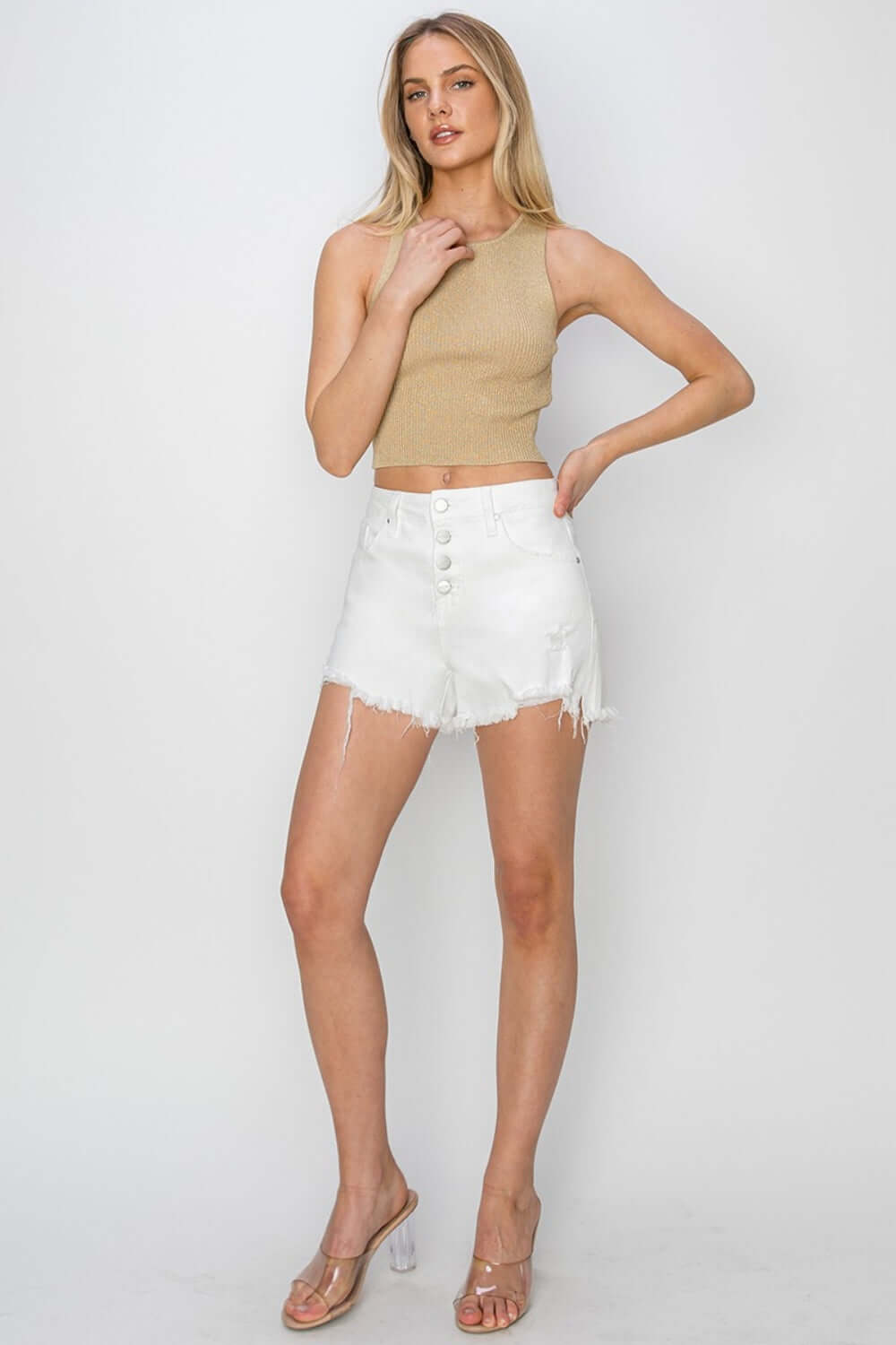 Model wearing Risen Jeans Button Fly Frayed Hem Denim Shorts in white with a beige cropped top and clear wedge sandals.