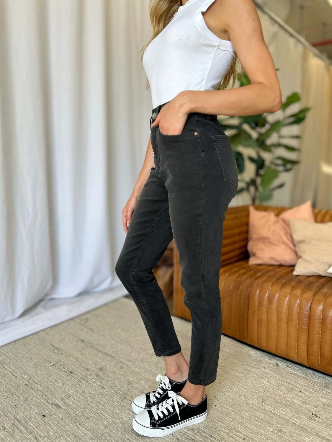 Woman wearing RFM Full Size High Rise Tummy Control Skinny Jeans in black, standing in a stylish room with a brown sofa and plant.