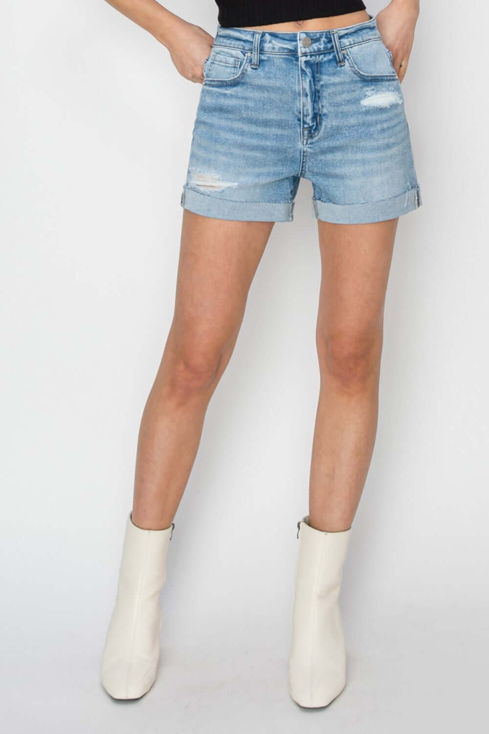 Woman modeling distressed mid-rise waist denim shorts from Risen Jeans, showcasing a trendy summer look with white ankle boots.