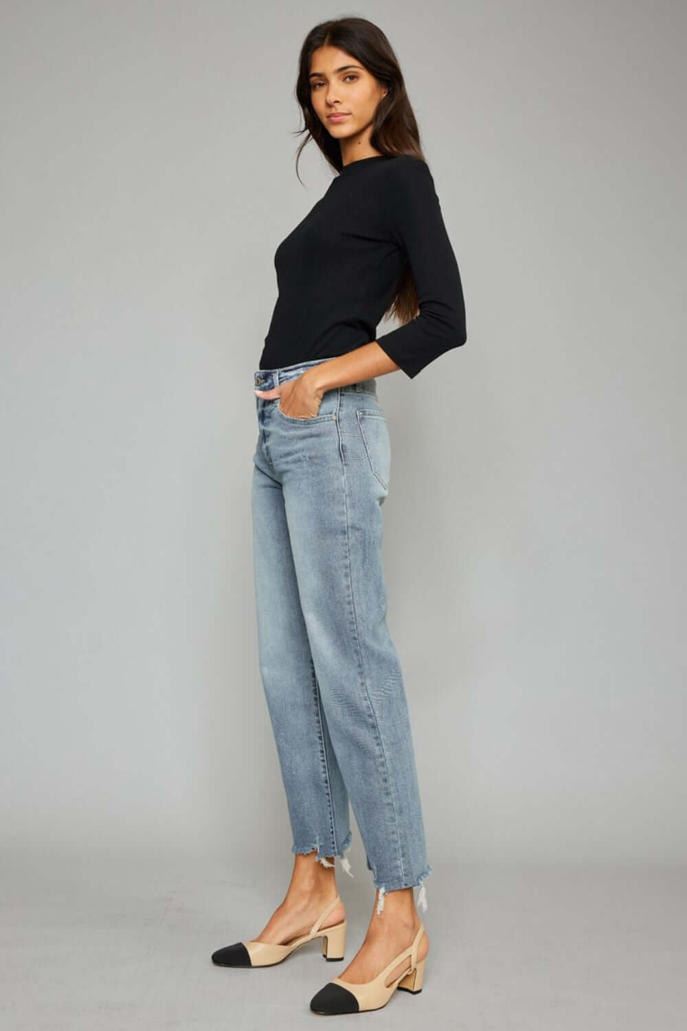 Woman wearing trendy high waist raw hem cropped wide leg jeans and black top standing against a gray background