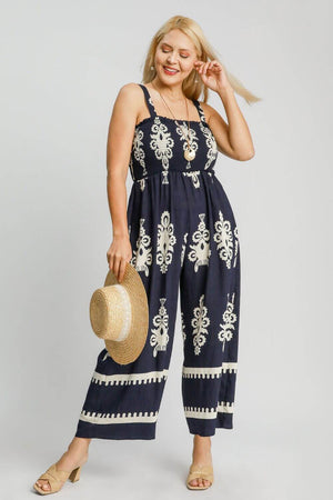 Plus size woman in navy smocked printed wide leg jumpsuit, holding a straw hat, perfect for summer outings.