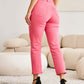 Woman wearing pink high-waist raw hem jeans showcasing tummy control and slender leg line, standing in a modern living room. RFM Jeans