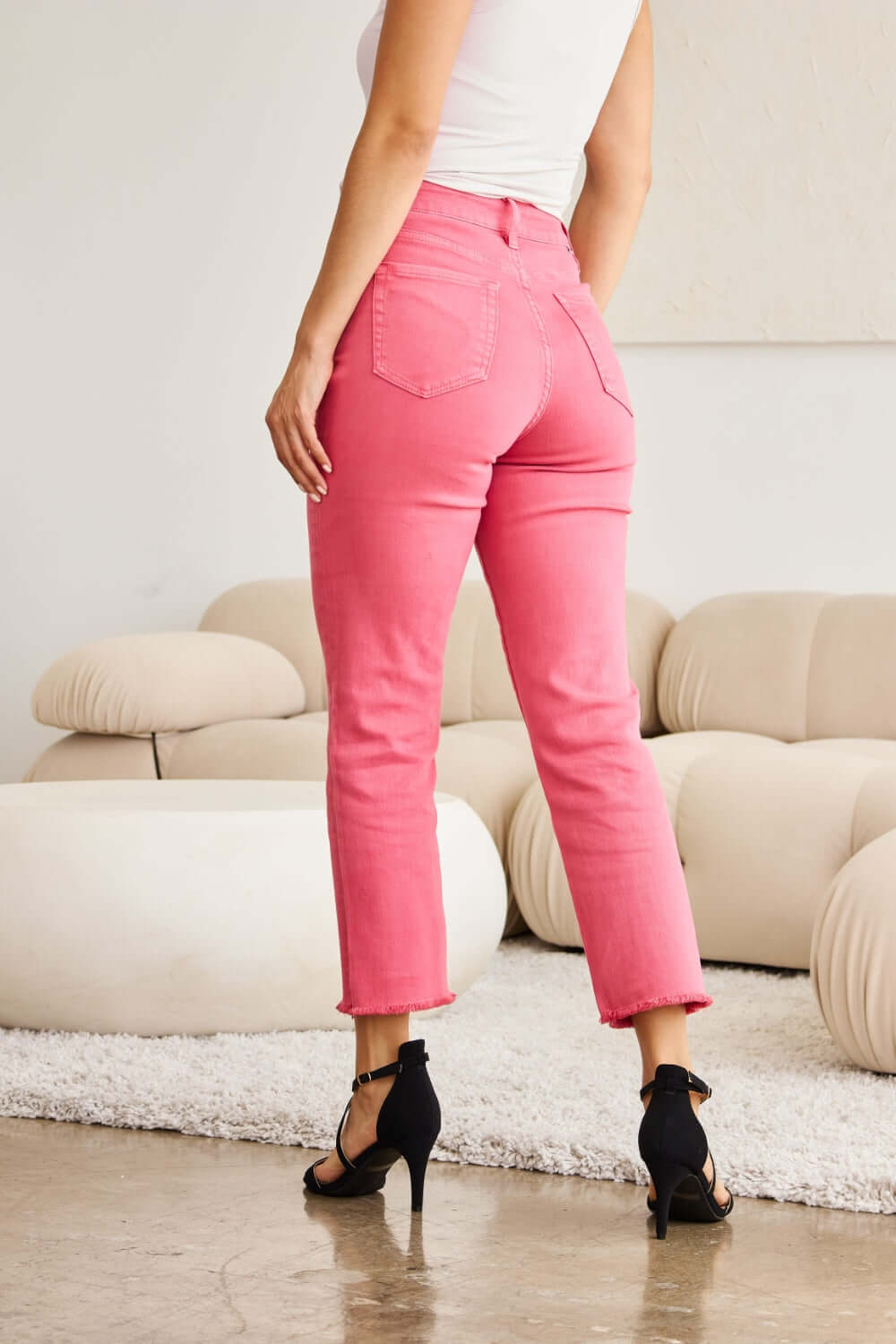 Woman wearing pink high-waist raw hem jeans showcasing tummy control and slender leg line, standing in a modern living room. RFM Jeans